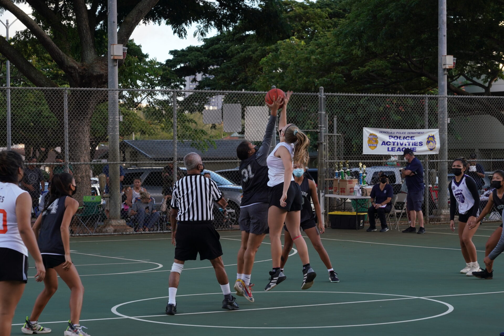 Two participants from the Police Activities League leap in the air on an outdoor basketball court, both reaching for the basketball to gain possession for their team.