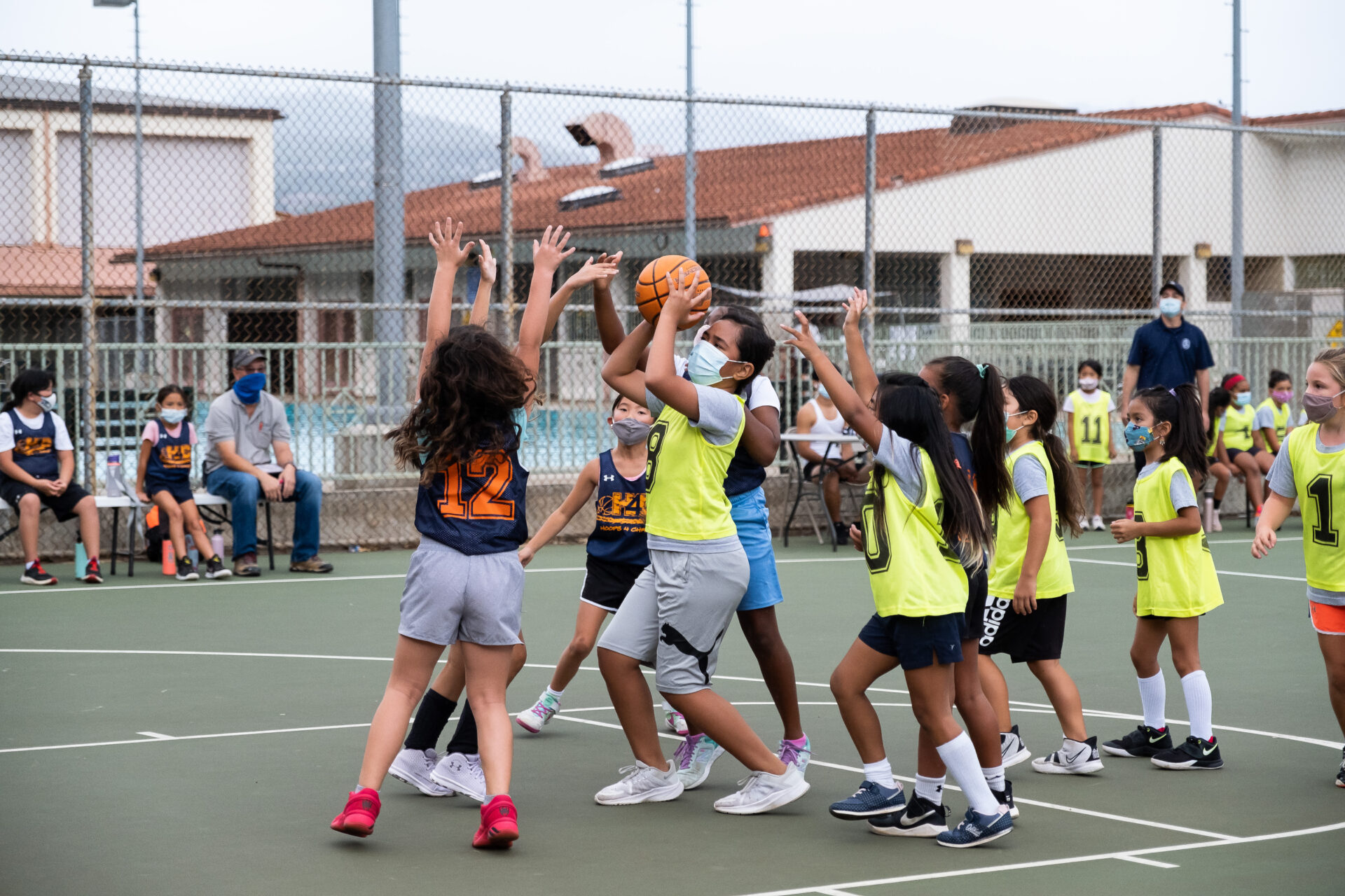 A young participant from the Police Activities League is on an outdoor basketball court, moving with the basketball and preparing to shoot it into the hoop. Other participants are nearby, with their hands raised, attempting to defend.