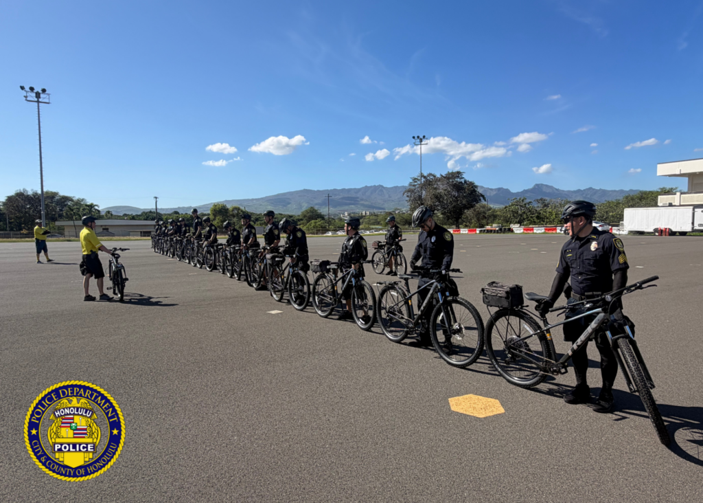 Photo of a large open paved area with more than a dozen Honolulu Police Officers attired in uniform, wearing a bicycle helmet, and standing next to a bicycle. There are two others whoc are the IPMBA (International Police Mountain Bike Association) Bicycle Instructors and are attired in a yellow polo shirt, shorts, and wearing bicycle helmets.