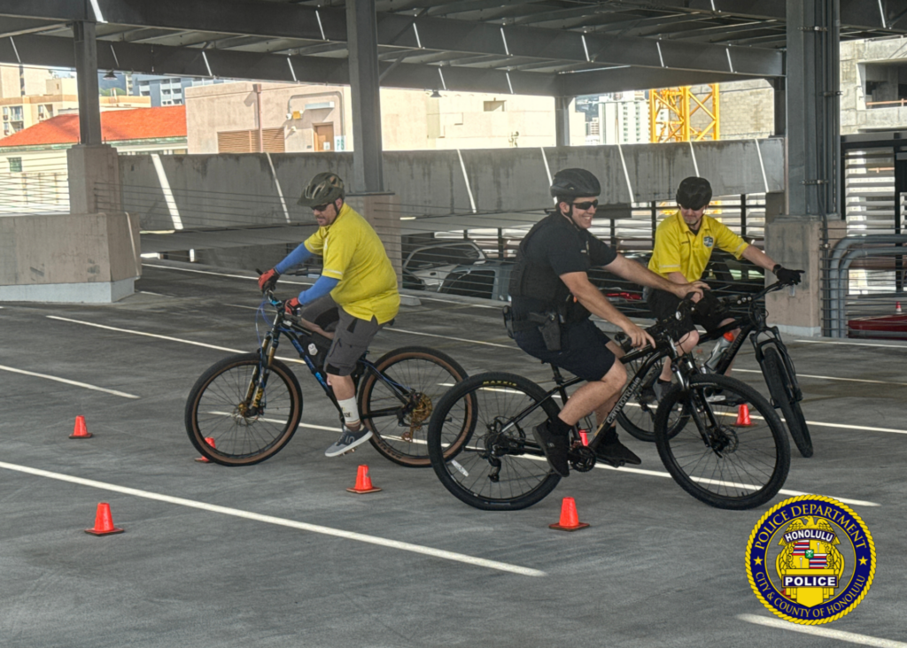 Photo with three individuals all seating and operating bicycles around small orange cones that are set up in a course. One individual is attired in a Honolulu Police uniform that is appropriate for riding a bicycle and a helmet. The other two individuals are IPMBA (International Police Mountain Bike Association) Bicycle Instructors and is also seated on bicycles and operating the course.