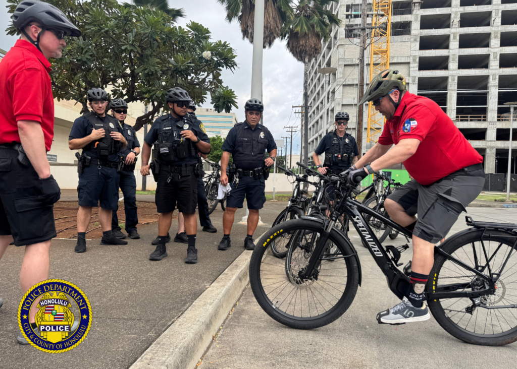 Photo of eight individuals, two of which are IPMBA (International Police Mountain Bike Association) Bicycle Instructors. One instructor is on a bicycle doing what appears to be a demonstration. The six other individuals are Honolulu Police officers attires in bicycle appropriate uniform attire and including bicycle helmets.