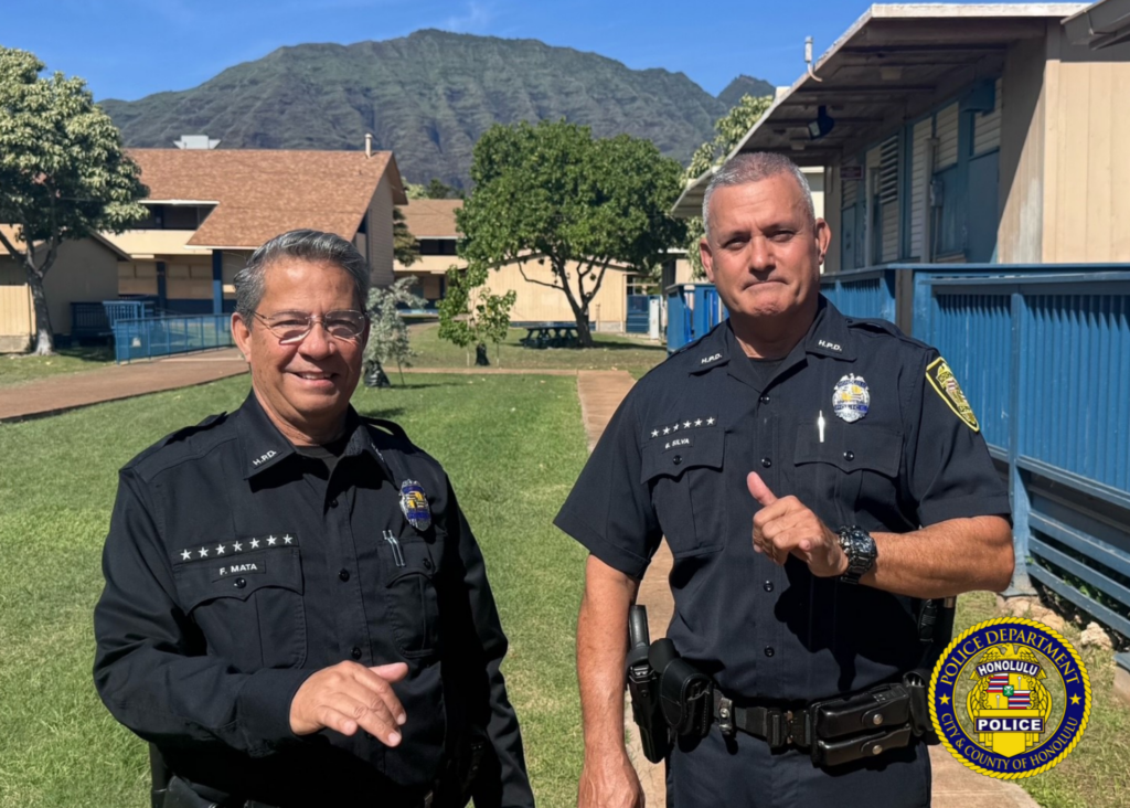 Half-body photo of two Honolulu Police Department School Resource Officers. Both are attired in their HPD uniforms and posing with a shaka. The background appears to be outdoors on campus with green grass and buildings.