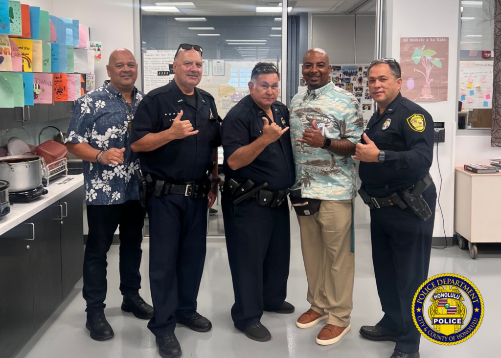 Full-body photo five males, three are Honolulu Police Department School Resource Officers. All officers are attired in their HPD uniforms. Two of the males are attired in an Aloha shirt and long pants. All males are posing with a shaka. Background setting is what appears to be a classroom.