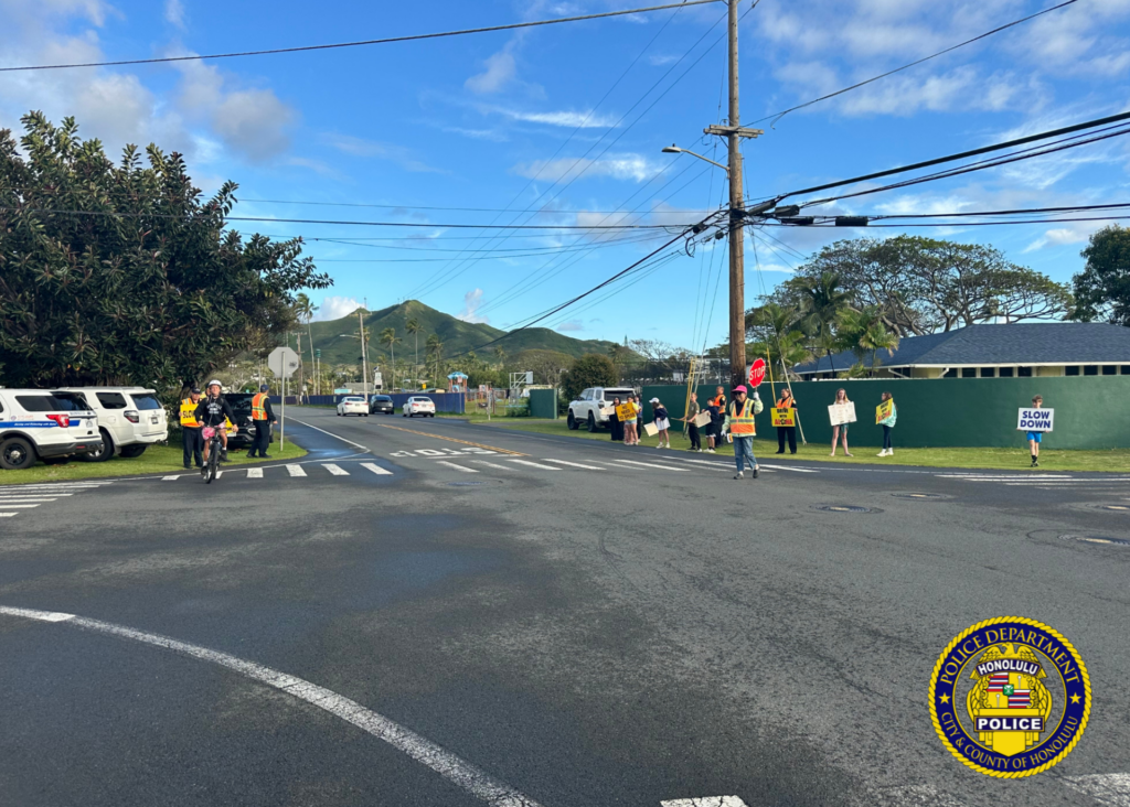 Police officers and students participate in a traffic safety sign-waving event near a school crosswalk as vehicles pass by.