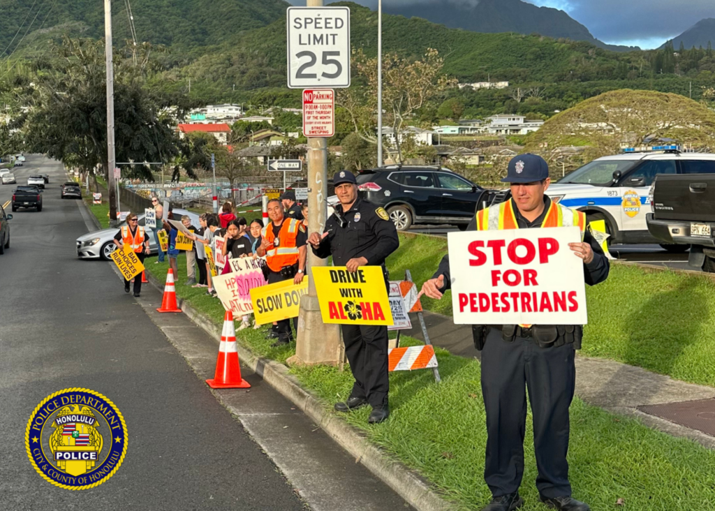HPD officers and elementary staff and students standing along the curb line waving various traffic safety messages to passing drivers.