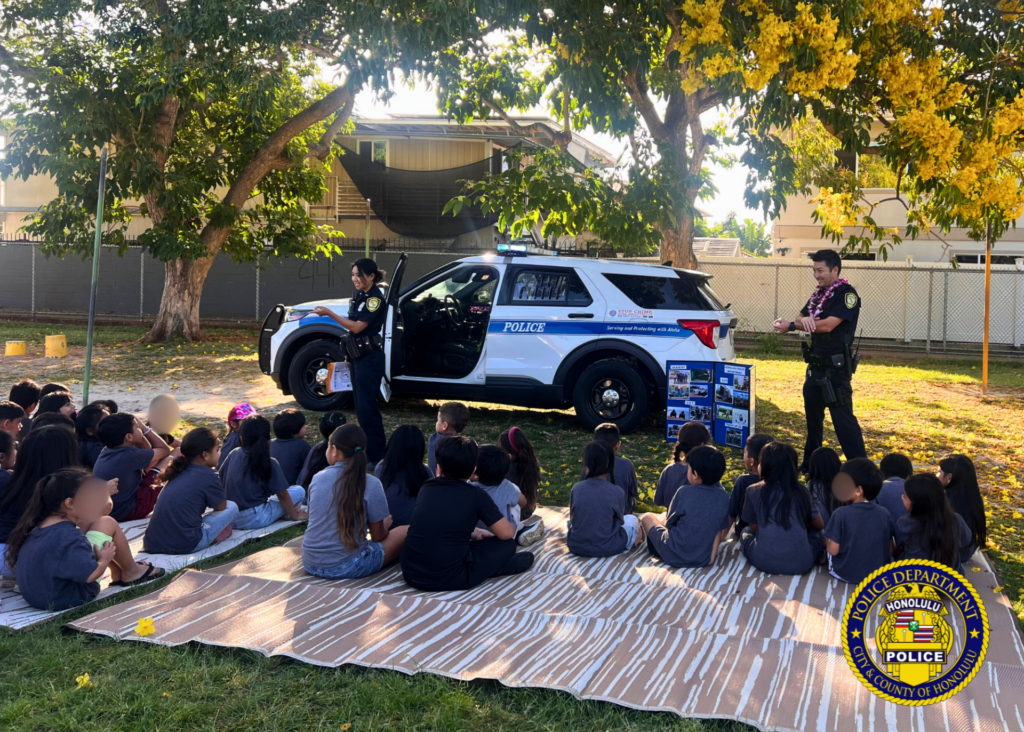HPD officers standing in front a group of children who are seated on mats on the grass