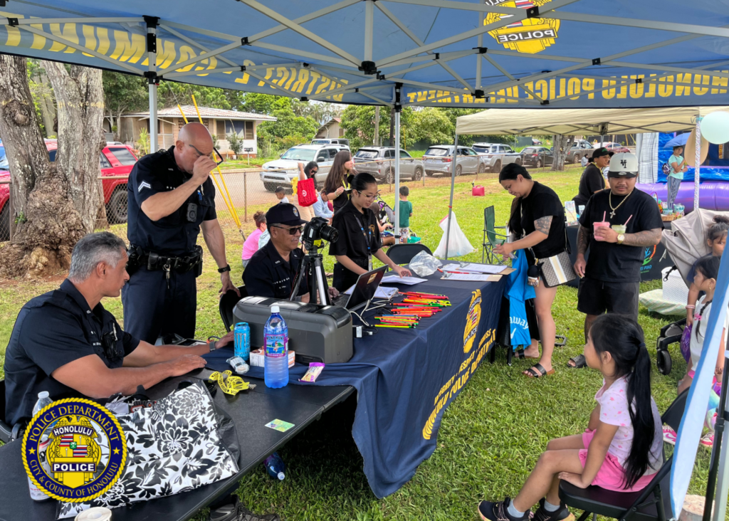 Honolulu Police Department officers assist a child with creating a keiki ID card at a community event booth, while families wait nearby under tents in a park setting.