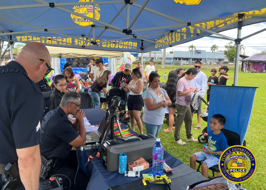 Honolulu Police Department officers assist a child with creating a keiki ID card at a community event booth, while families wait nearby under tents in a park setting.