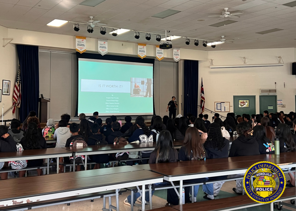 A Honolulu Police Department officer speaks to students while a presentation is displayed on a projector.