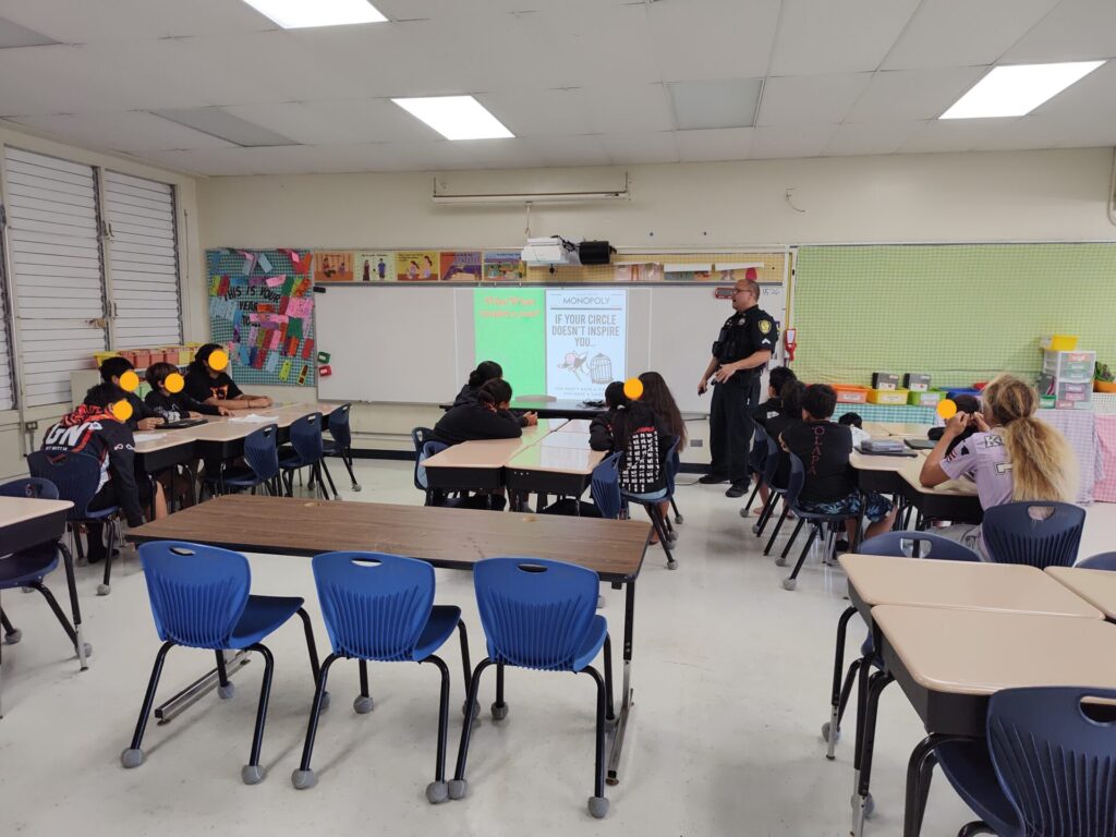 Honolulu police officer in uniform standing at the front of a classroom, engaging students during a presentation.