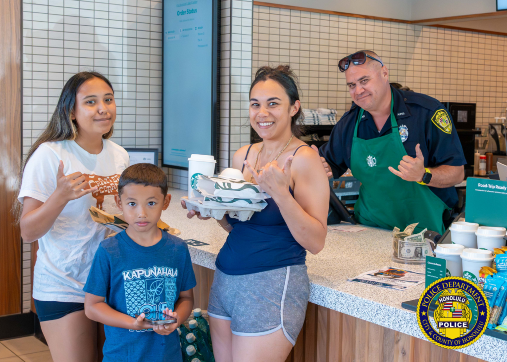 Inside a Starbucks, a police officer wearing a green Starbucks apron stands behind the counter with three customers, including two children. The customers hold drinks and a tray of beverages while smiling toward the camera and making shaka hand gestures. Starbucks cups and merchandise are visible on the counter. Honolulu Police Department logo watermark appears in the lower-right corner.