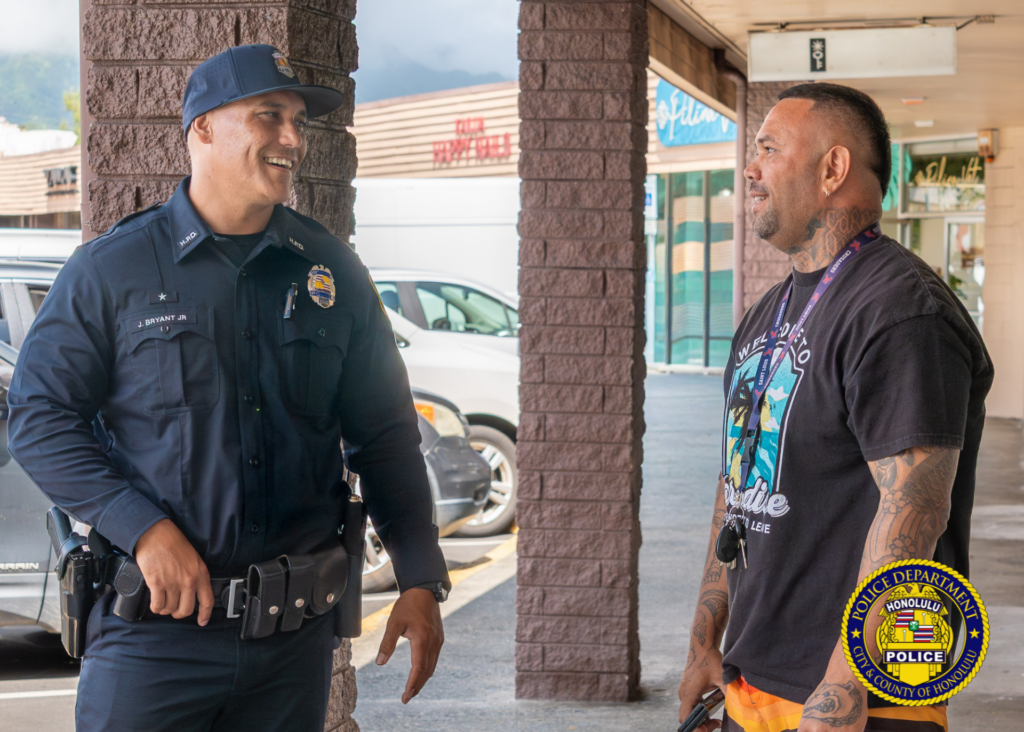 A uniformed police officer and a community member stand under a shopping center walkway engaged in conversation. The officer smiles while facing the community member, who has visible arm and neck tattoos. Parked vehicles and storefronts are visible in the background. Honolulu Police Department logo watermark appears in the lower-right corner.