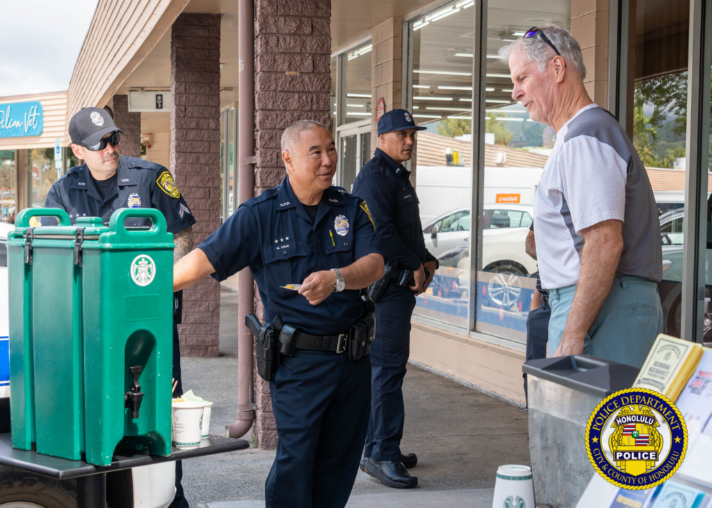 Three uniformed police officers speak with a community member outside a shopping center. One officer gestures while talking. Large green beverage dispensers sit on a table in the foreground, and storefront windows line the walkway behind them. Honolulu Police Department logo watermark appears in the lower-right corner.