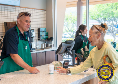 Inside a Starbucks, a police officer wearing a green Starbucks apron stands behind the counter speaking with a customer who is handing over cash. Another Starbucks employee stands at a register in the background. A coffee cup sits on the counter between them. Honolulu Police Department logo watermark appears in the lower-right corner.
