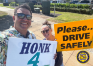 Two people stand on a sidewalk holding traffic-safety signs and smiling toward the camera. One sign reads “HONK 4 SAFETY!” and the other reads “Please… DRIVE SAFELY.” Vehicles, landscaping, and utility boxes are visible in the background. Honolulu Police Department logo watermark appears in the lower-right corner. Visible text: HONK 4 SAFETY! Please… DRIVE SAFELY