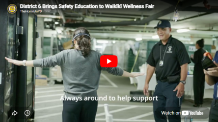 Video thumbnail showing a person standing with arms extended while interacting with a touchscreen display in an indoor parking-garage setting. A uniformed public safety officer stands nearby watching and smiling. White on-screen text at the top reads, “District 6 Brings Safety Education to Waikīkī Wellness Fair,” with “TheHonoluluPD” below it. White caption text near the bottom reads, “Always around to help support.” A red YouTube play button appears in the center, and a “Watch on YouTube” button appears in the lower-right corner.