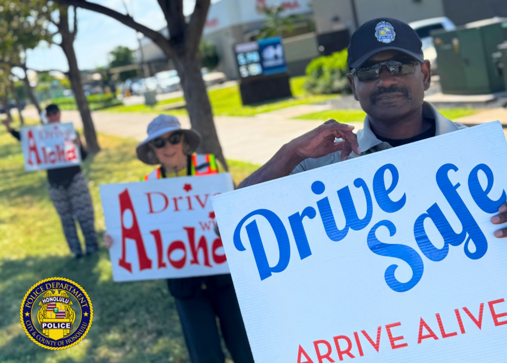 A person wearing a Honolulu Police cap stands on a grassy roadside holding a sign that reads “Drive Safe ARRIVE ALIVE.” Two additional people stand behind them holding traffic-safety signs, including one that reads “Drive with Aloha.” Trees, a sidewalk, and a roadway are visible in the background. Honolulu Police Department logo watermark appears in the lower-left corner. Visible text: Drive Safe ARRIVE ALIVE Drive with Aloha