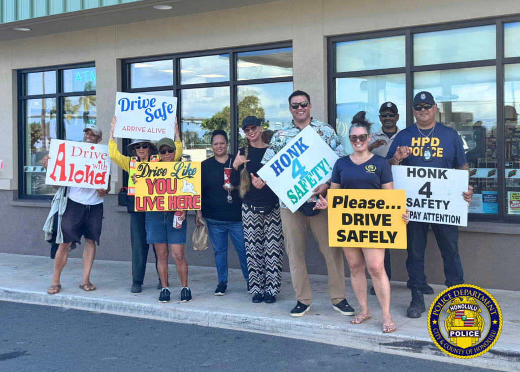 A group of community members and Honolulu Police personnel stand outside a storefront holding traffic-safety signs and smiling toward the camera. Signs promote safe driving, paying attention while driving, and driving with aloha. Honolulu Police Department logo watermark appears in the lower-right corner. Visible text: Drive with Aloha Drive Safe ARRIVE ALIVE Drive Like YOU LIVE HERE HONK 4 SAFETY! Please… DRIVE SAFELY HONK 4 SAFETY PAY ATTENTION