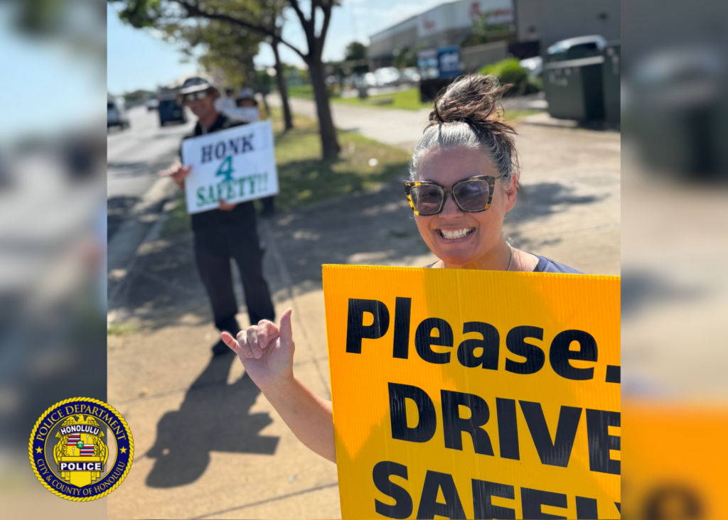 A person standing on a sidewalk smiles while holding a yellow traffic-safety sign that reads “Please… DRIVE SAFELY.” Another person stands farther back holding a sign that reads “HONK 4 SAFETY!” Trees, a roadway, and passing vehicles are visible in the background. Honolulu Police Department logo watermark appears in the lower-left corner. Visible text: Please… DRIVE SAFELY HONK 4 SAFETY!