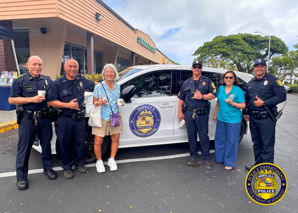 Six people stand beside a white Honolulu Police community policing vehicle in a shopping center parking lot. Four uniformed police officers and two community members smile toward the camera, several making shaka hand gestures. A Starbucks storefront is visible in the background. The vehicle door displays a circular “District 4 Honolulu Police Community Policing” emblem. Honolulu Police Department logo watermark appears in the lower-right corner.