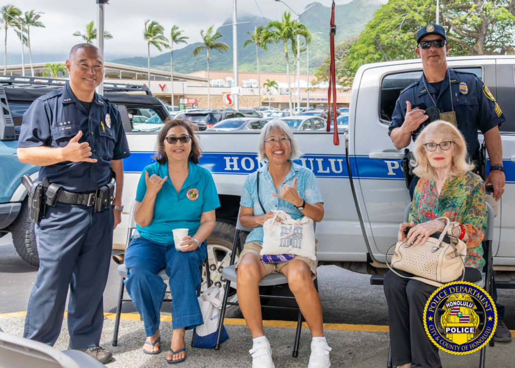 Two uniformed police officers and three community members pose in front of a Honolulu Police pickup truck. Two people are seated in folding chairs while others stand nearby. Several people make shaka hand gestures. Palm trees, parked vehicles, and mountains are visible in the background. Honolulu Police Department logo watermark appears in the lower-right corner.