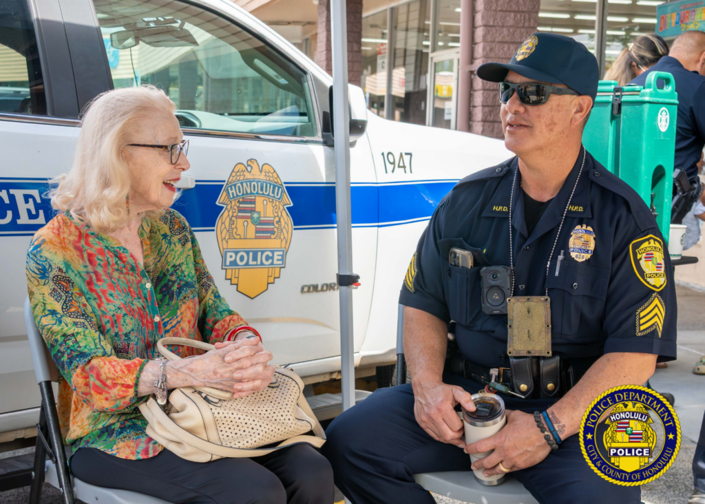 A seated community member and a uniformed police officer sit facing each other and talk beside a Honolulu Police pickup truck. The officer holds a travel mug while the community member holds a handbag on their lap. The Honolulu Police badge graphic is visible on the truck door behind them. Honolulu Police Department logo watermark appears in the lower-right corner.