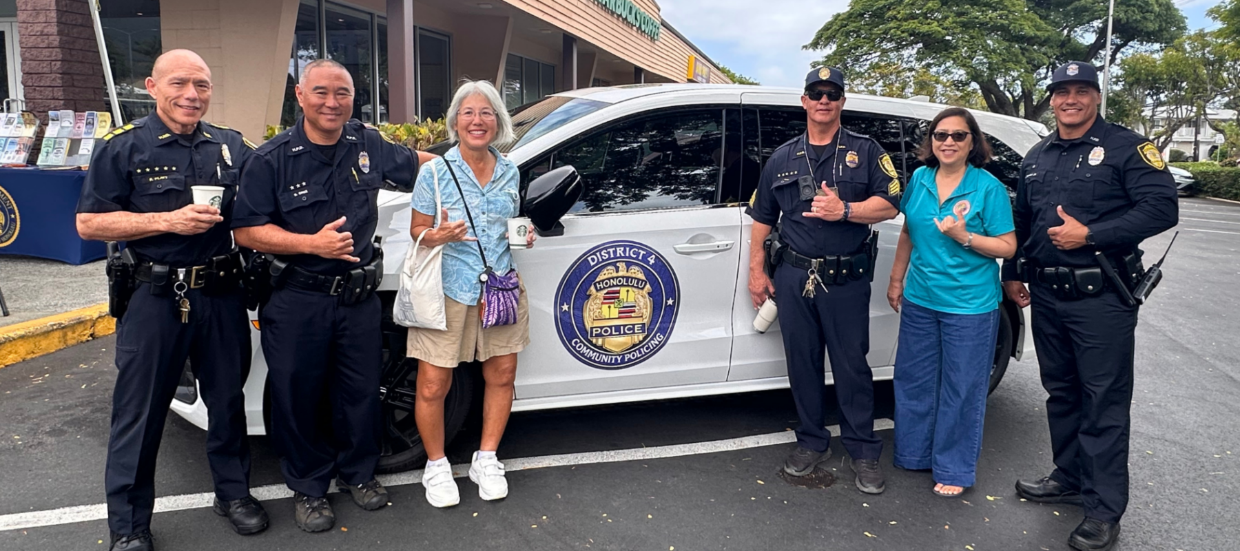 Six people stand beside a white Honolulu Police community policing vehicle in a shopping center parking lot. Four uniformed police officers and two community members smile toward the camera, several making shaka hand gestures. A Starbucks storefront is visible in the background. The vehicle door displays a circular “District 4 Honolulu Police Community Policing” emblem.