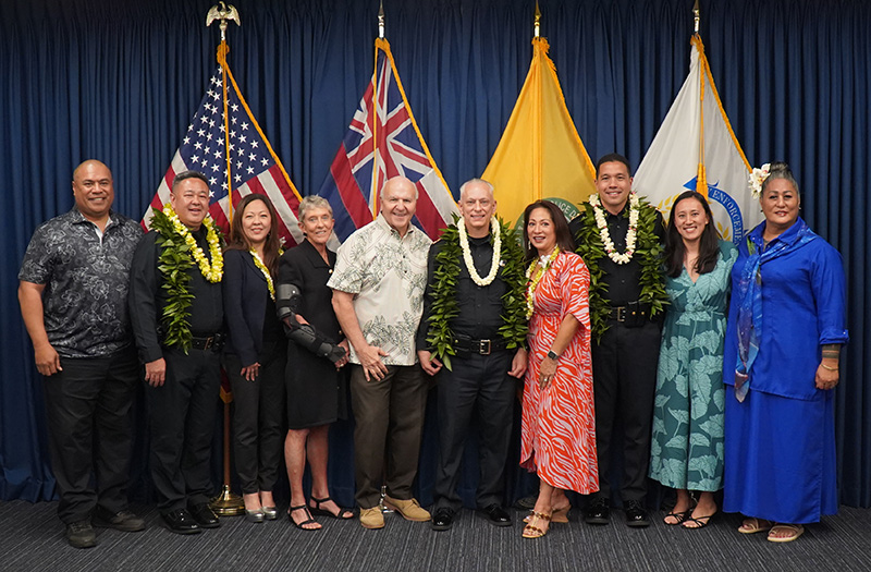 A group photo of Chief David Lazar, Deputy Chief Brandon Nakasato, Deputy Chief Parker Bode, their friends and family, and Mayor Rick Blangiardi in front of the flag display and a blue curtain background.